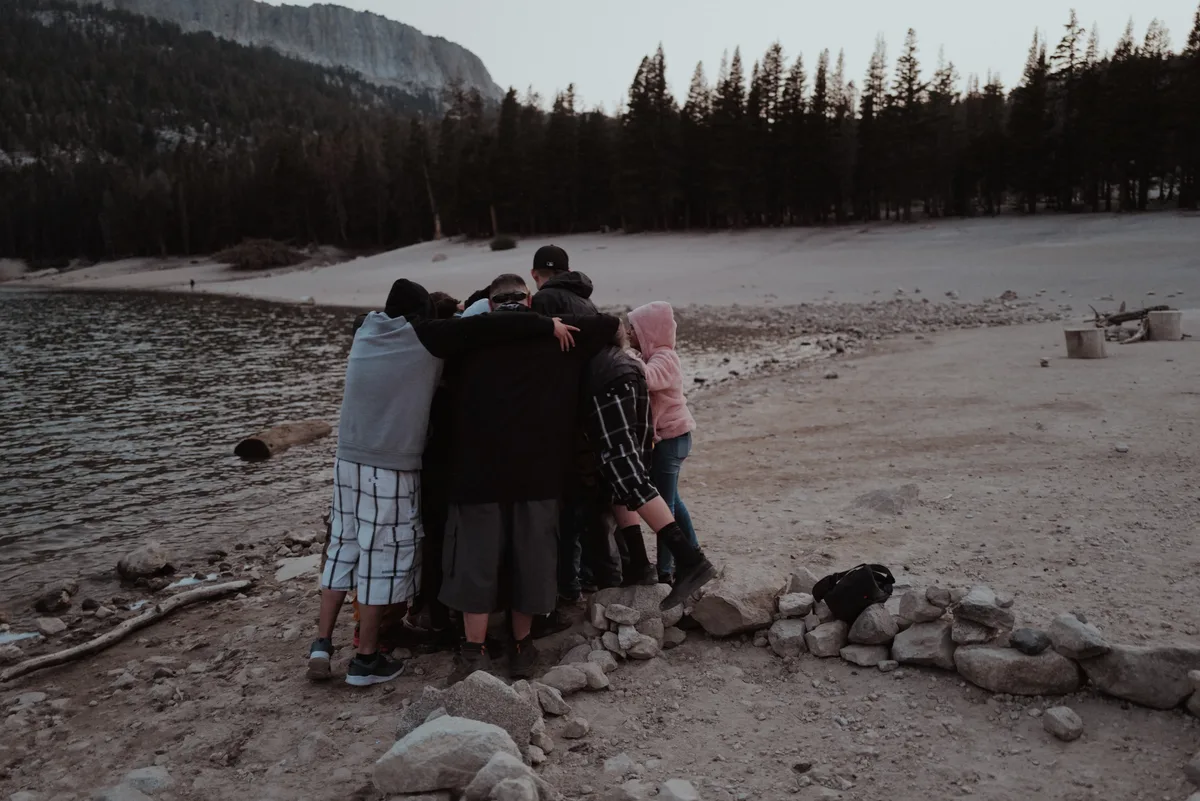 A group stands with arms around each other beside a lake.